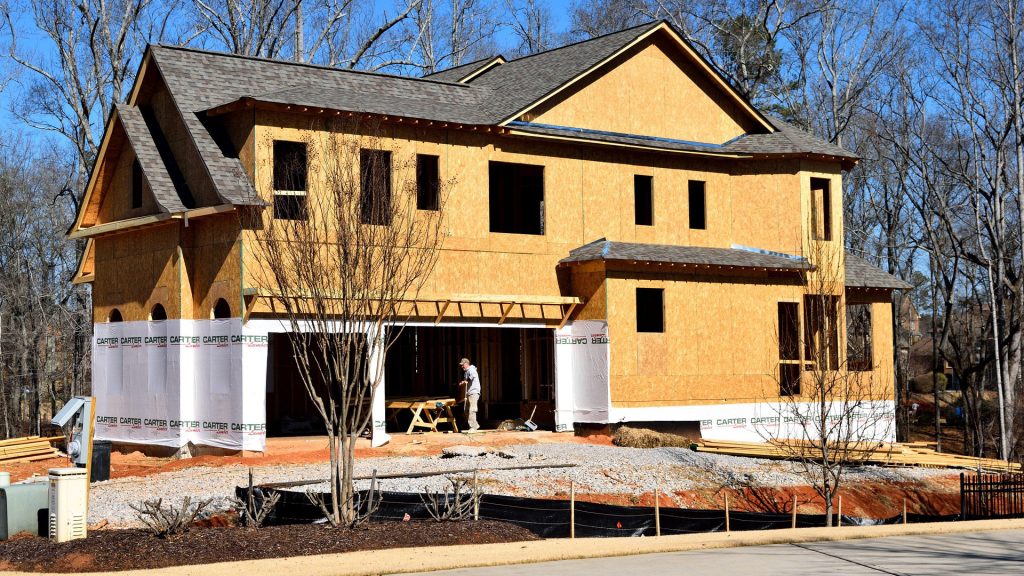 A two-story home under construction with exposed plywood framing and roof shingles installed. A worker is preparing materials inside the open garage. The site features gravel groundwork and construction wrap labeled "CARTER" around the lower exterior. Leafless trees and a clear blue sky frame the background.