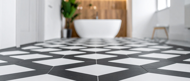 Close-up view of a modern bathroom with radiant heated black and white geometric tile flooring, highlighting the sleek pattern and even warmth; a freestanding tub, wood accent wall, and single green plant add contrast and texture in the background.